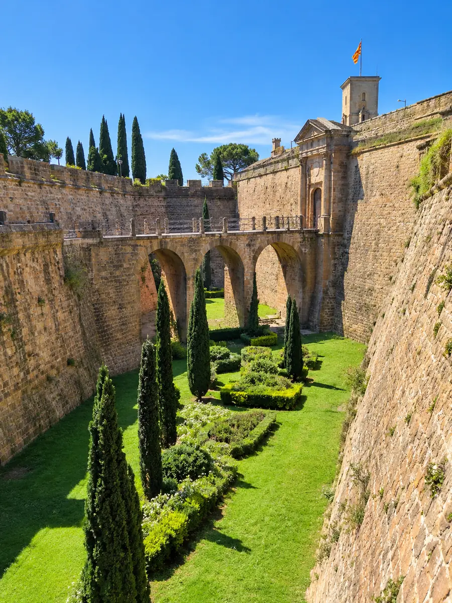 Der grüne Garten im Burggraben des Kastells von Montjuïc zwischen geböschten Steinmauern, eine Bogenbrücke führt zum Tor