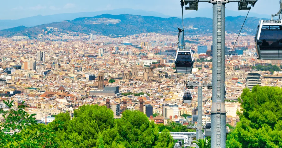 Seilbahngondeln und ein Mast über grünen Pinien, dahinter ganz Barcelona und die Hügel der Collserola
